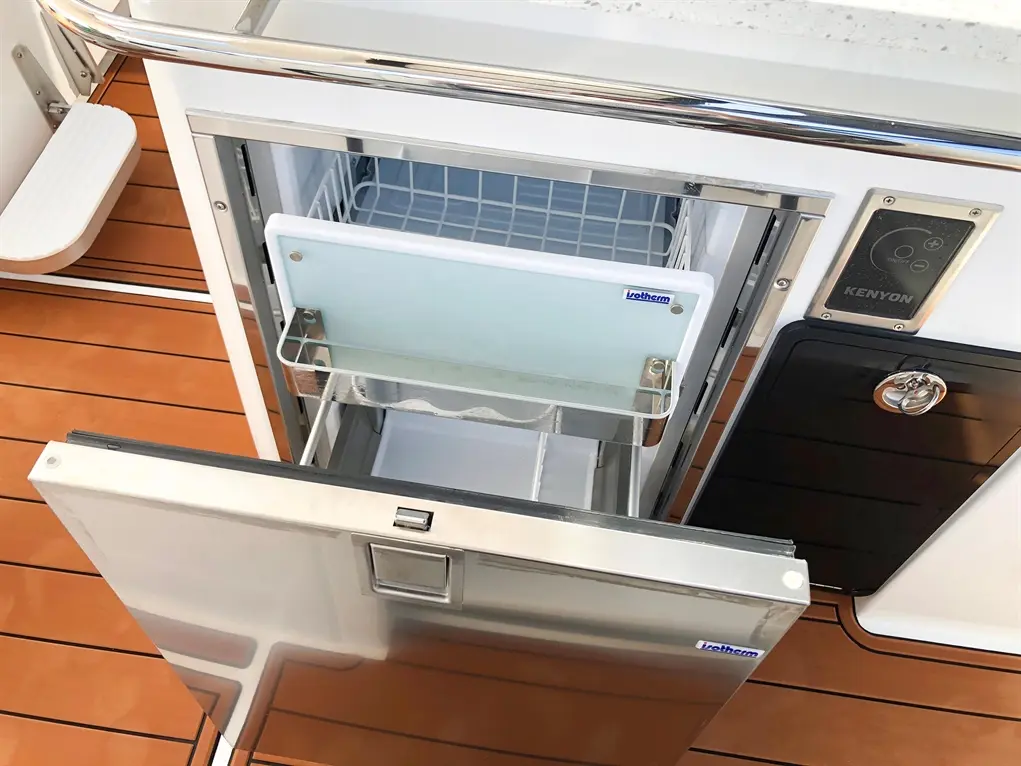 A stainless steel marine fridge with a drawer open, featuring a Kenyon control panel and a wooden deck background.